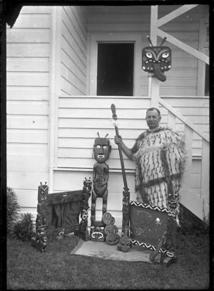 Albert Percy Godber wearing a Maori cloak, holding a taiaha, standing beside a collection of Maori carvings, including two fire-screens, which he carved