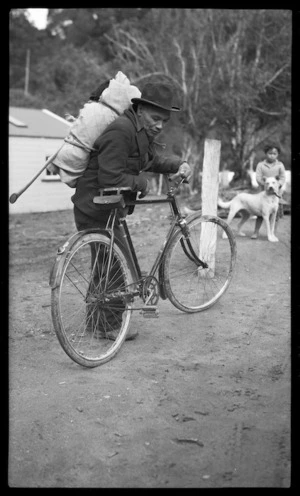 Maori man with a swag on his back leaning against a bicycle