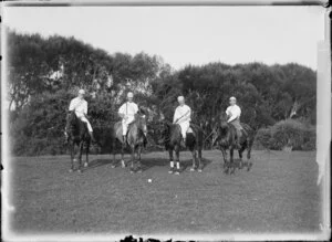 Four identified men on horseback and dressed for a game of polo and holding polo mallets, with a polo ball on the ground in front of the group, location unknown