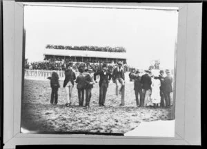 Group of men standing in front of a crowded grandstand on race day, Wanganui racecourse