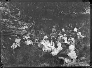 An unidentified group of finely dressed men, women, and children, at a picnic, posing on a grassy bank under trees, location unknown
