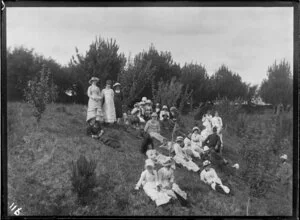 An unidentified group on a picnic recline on a bank, with three upstanding, including a policeman, in an unknown garden