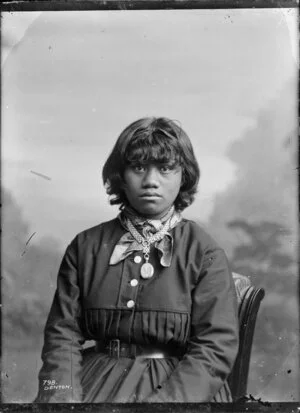 Studio portrait of Maori girl wearing dark pleated dress with pendant on woven chain(?) worn over scarf tied at neck, probably from the Whanganui region. Photograph taken by Frank J Denton
