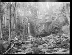 Four women and a man beside a rock-strewn stream with a steep cliff and waterfall behind, Australia