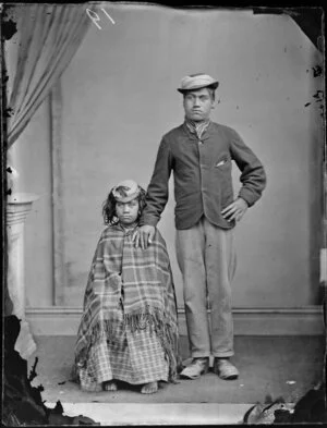 Young Maori couple, the man in a coat trousers and cravat, the woman with a dress and blanket, both with hats
