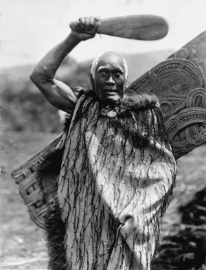 Unidentified elderly Maori man, wearing a tag cloak and wielding a patu