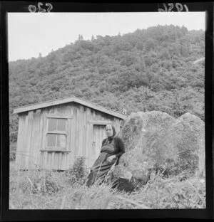 Unidentified woman, sitting on a rock outside a small wooden dwelling, Waihi Village, Lake Taupo