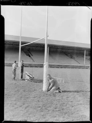 Mr Meates, groundskeeper at Athletic Park, Wellington, tying protective pads on the goalposts with the help of two unidentified men