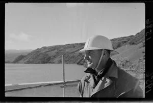 An unidentified worker models a helmet worn by workers on the hydroelectric project at Roxburgh, including dam under construction and the Clutha River in the background