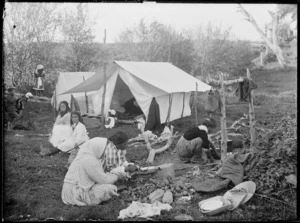 Maori women and girls by tents, preparing food, location unidentified; peeling potatoes in foreground