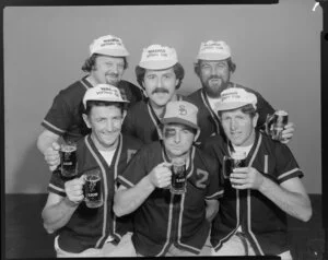 People (including one with fake black eye) in softball uniforms drinking Lion beer in studio