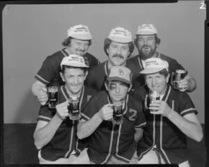 People (including one with fake black eye) in softball uniforms drinking Lion beer in studio