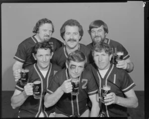 People (including one with fake black eye) in softball uniforms drinking Lion beer in studio