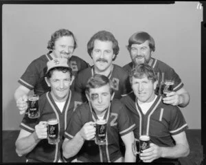 People (including one with fake black eye) in softball uniforms drinking Lion beer in studio