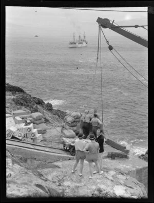 Group of people and supplies beside winch with coastal steamer in the distance