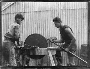 Two Māori men sharpening an implement on a manual grinding stone