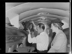 Airline steward F Bennett stowing luggage in an overhead storage compartment in a Dakota aircraft