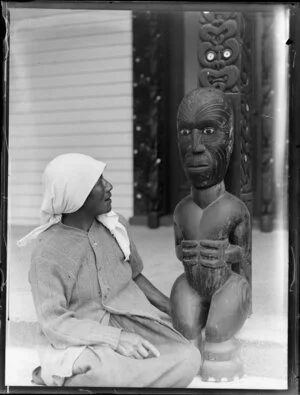 Maori wahine sitting next to a Maori sculpture, location unidentified