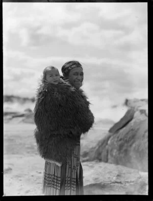 Maori woman carrying a young child on her back wrapped in a feather cloak