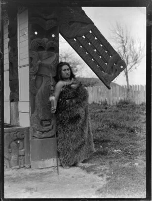 Maori woman dressed in a feather cloak, holding a taiaha, location unidentified