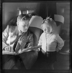 Young child Ms Shandler, a passenger on National Airways Corporation Douglas DC-3 flight from Auckland to Fiji, reads a newspaper with an unidentified male passenger