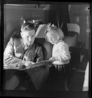 Young child Ms Shandler, a passenger on National Airways Corporation Douglas DC-3 flight from Auckland to Fiji, reading a newspaper with an unidentified male passenger
