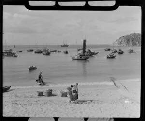 Chinese fishing boats or sampans beside a sandy beach; two men carry baskets of fish up the beach, unidentified location, Hong Kong