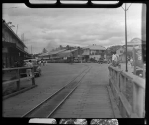 Street in Ba, Fiji, showing railway tracks with a sugar cane train on it in the centre of the road, Goverdhanbhai General Merchants and other shops, vehicles and people