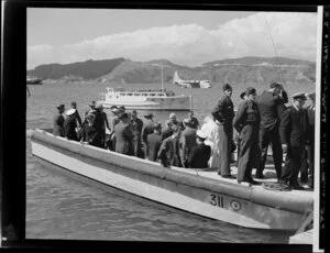 Unidentified passengers and Tasman Empire Airways Limited crew disembark from a boat at the flying boat base near Hataitai Beach, Wellington, with the Short S.45 Solent flying boat, R.M.A Araragi (ZK-AMM), moored in Evans Bay in the background