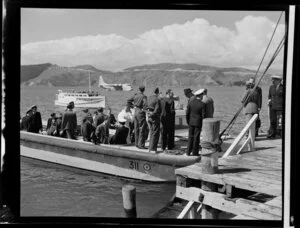 Unidentified passengers and Tasman Empire Airways Limited crew arrive on a boat at the the flying boat base near Hataitai Beach, Wellington, with the Short S.45 Solent flying boat, R.M.A Araragi (ZK-AMM), moored in Evans Bay in the background