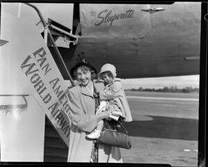 Passenger Mrs Holloway (holding her young child), having arrived on a Pan American World Airways flight, stands in front of Douglas DC-4 Clipper Westward Ho (N88948) at Whenuapai aerodrome, Waitakere City, Auckland