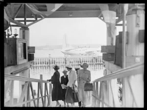 Unidentified group of women walking up a gangplank [at a Tasman Empire Airways Ltd event]; seaplane ZK-AMM is behind