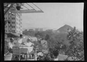 K Gibson and R Sayers (left to right) sit at a table in a patio and are served tea by a local waiter, [Victoria Peak?] Hong Kong, including apartment buildings in background
