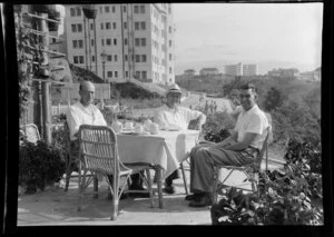 K Gibson, R Sayers and Leo Lemuel White (left to right) take tea on a patio, [Mt Victoria?] Hong Kong, including apartment buildings in background