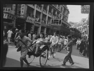 Busy street scene, Hong Kong, including a woman riding in a hand drawn rickshaw and double-decker trams