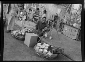 Market scene, Hong Kong, featuring man selling kitchenware from baskets joined by a shoulder yoke
