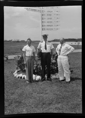 From left are Leo White, Captain J C Pollock, Captain F Ambrose, standing in front of a Qantas Empire Airways signpost showing directions to overseas airports, Tengah, Singapore