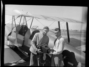 C Stewart [pilot?] and photographer Leo Lemuel White (left to right) holding a field camera in front of an airplane, Mangere Aerodrome, Manukau City, Auckland Region