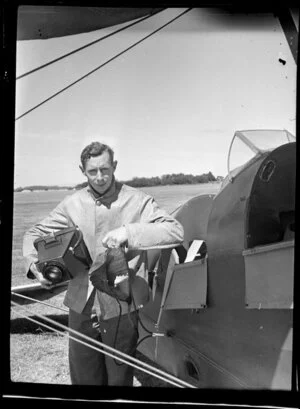C Stewart [pilot?], who is holding a field camera, alongside airplane, Mangere Aerodrome, Manukau City, Auckland Region