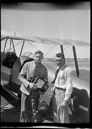 C Stewart [pilot?], who is holding a field camera, and photographer Leo Lemuel White, in front of a plane, Mangere Aerodrome, Manukau City, Auckland Region