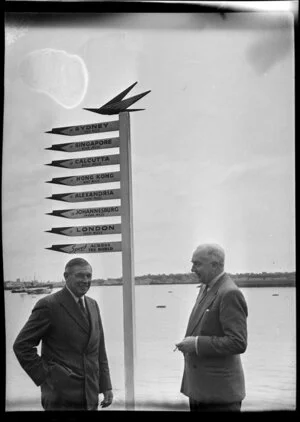 British Overseas Airways Corporation Chairman Sir Miles Thomas and Tasman Empire Airways Chairman Sir Leonard Isitt, beside a sign at the Auckland flying boat base, listing destinations and their distance from Auckland (including Syndey, Singapore, Calcutta, Hong Kong, Alexandria, Johannesburg and London)