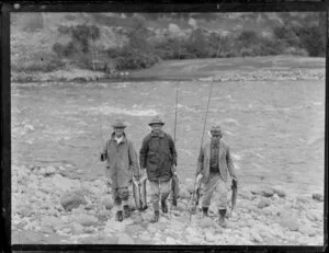 Three unidentified fishermen, all of whom wear hats, on a stony riverbank with their rods and catch of trout