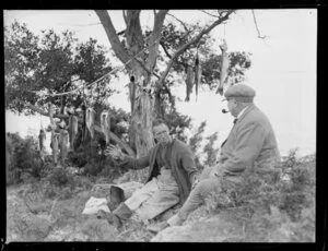 Two unidentified fishermen smoking on bank, with gutted trout drying in tree