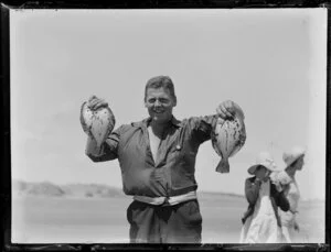 Unidentified man [fisherman?] holding up fish, Ninety Mile Beach, Far North District, Northland Region, with unidentified women in background