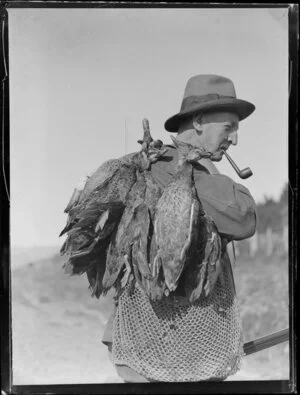 Duck shooting, featuring unidentified hunter, who is wearing a hat and smoking a pipe, carrlying dead ducks and gun, location unknown