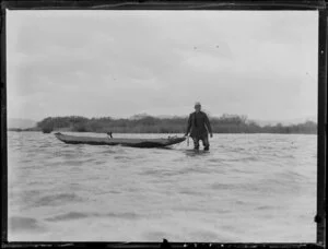 Duck shooting, featuring hunter wading through water towing boat, in which sits a small dog [border collie?], location unknown