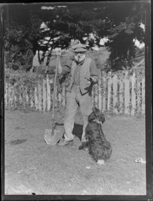 Unidentified elderly man, who is smoking a pipe and wearing a knitted hat, with dog and shovel, location unknown