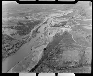 View down Clutha River from Roxburgh Dam site, Central Otago, showing excavation of diversion channel with heavy earth moving machinery, workers accommodation and site buildings and road down valley beyond