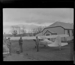 Auster aircraft ZK-AWS outside the Omarama Memorial Hall with Harry Wigley between two unidentified men, Waitaki County