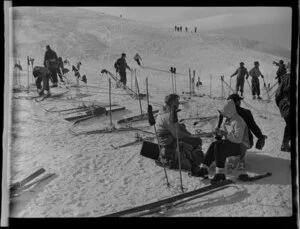 Unidentified man and woman sitting on stumps in the snow surrounded by skis, Coronet Peak, Otago